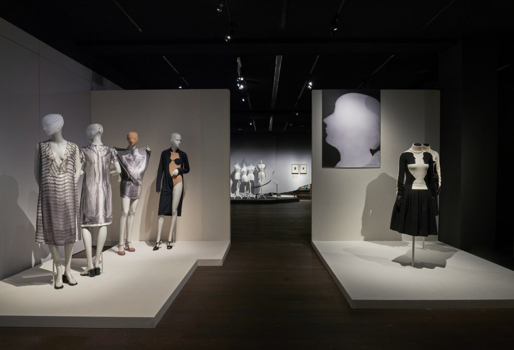Gallery view showing two plinths featuring four mannequins wearing monochrome dresses on the left' on the right two tailors' dummies featuring Surrealist-inspired dresses. In the background is another gallery featuring a group of tailors' dummies.