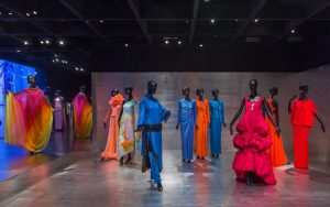 12 dresses, ranging from blue, pink and orange and grouped into pairs, are worn on expressive mannequins on a brushed metal floor and wall.