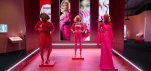 View of a pink gallery with three mannequins wearing pink outfits. In the background are close-up projections of Marilyn Monroe, Winnie Harlow and Rihanna.