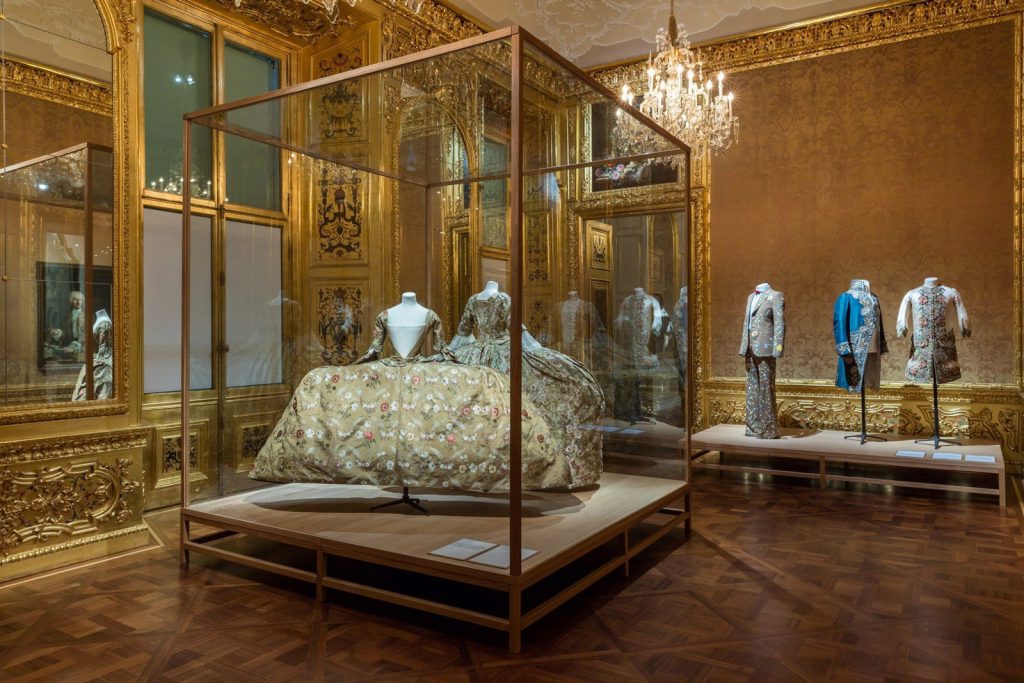 Two panniered skirts are displayed in a brown wooden frame in a gilt gold room. Three embroidered menswear outfits are displayed in the background.