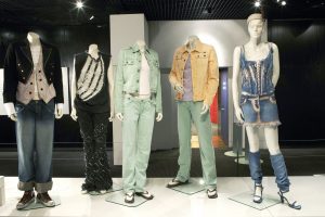 Five outfits are worn on white mannequins; the mannequin on the right has a head. The far-left outfit is a black bolero-style jacket and patterned waistcoat, white shirt, jeans and Converses.; the second is a sleeveless black distressed top and embroidered black skirt. The middle outfit is a light blue denim jacket, white top and matching denim jeans; on his right is a washed yellow jacket, mauve top and blue jeans. The last mannequin wearing a pill-box hat, denim corset, acid-washed mini-skirt, denim leg-warmers and blue heels.