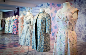 Two dresses and a coat are worn on three white tailor's dummy's in front of a wall with varying print patterns. More dresses are on display in the background.