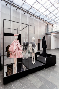 A pink dress with a matching draped cape and fascinator stands next to a 3D printed honeycomb-patterned dress and a black dress in front a black framed structure.