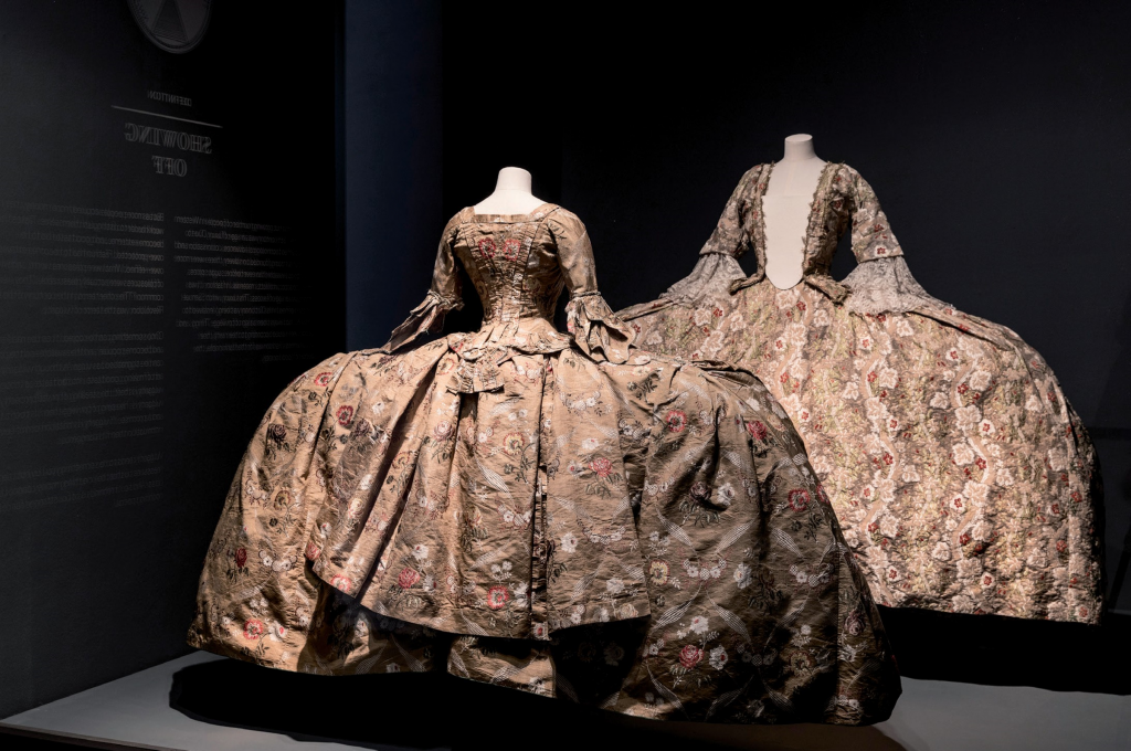 Two dresses with panniers are displayed in front of a dark background