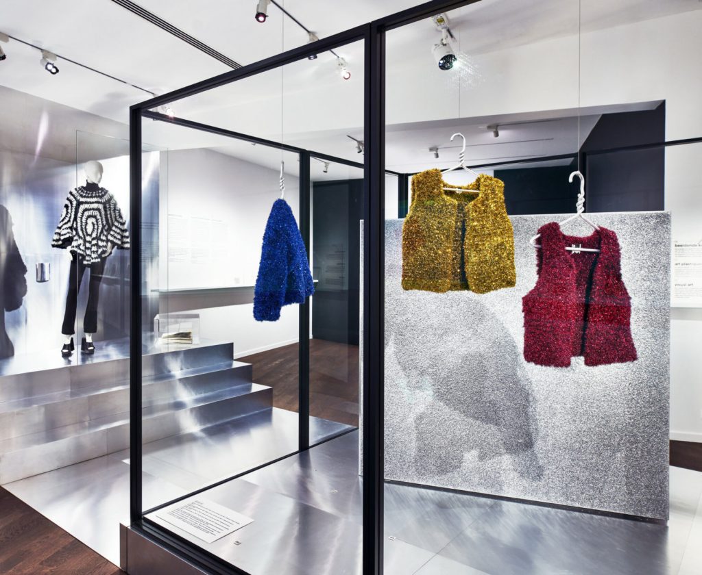 Three sparkly lurex jackets and waistcoats are hung on hangers on a silver framed structure. The jacket is blue and the waistcoats are gold are red, respectively.