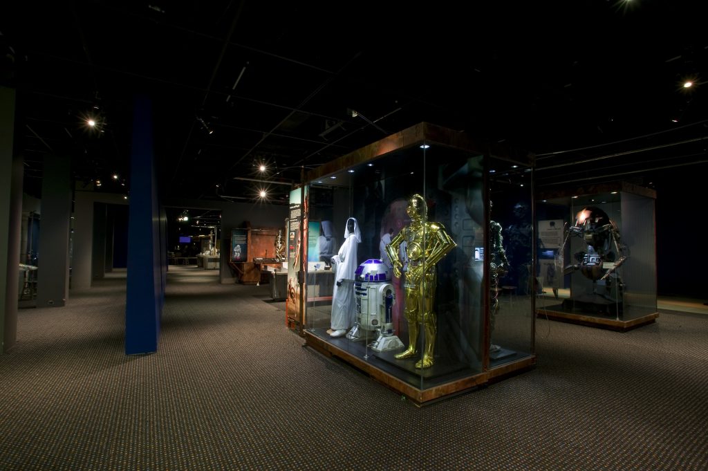 A display case shows costumes from Star Wars, including a white billowing gown (worn by Princess Leia), C3PO and R2-D2