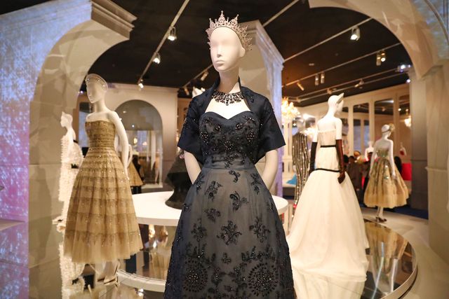 A mannequin wearing a tiara and black ballgown with a black bolero jacket and necklace. More dresses are displayed in the background.