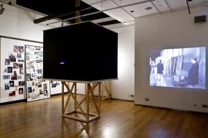 View of the gallery, showing a film projected onto a wall, a black box with a cut-out to view inside, and a wall of photographs