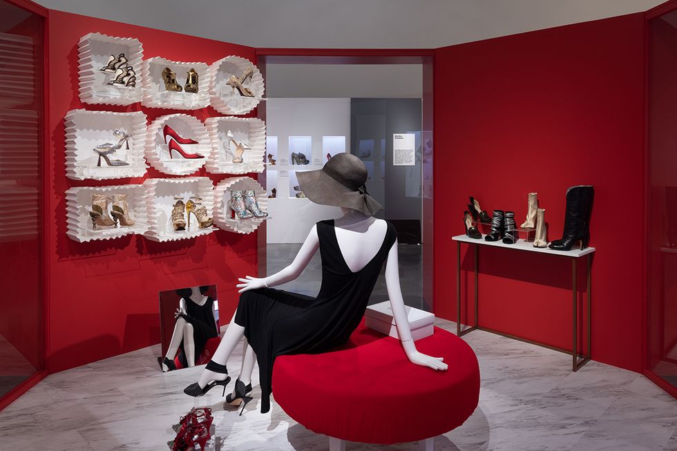 A sitting mannequin in front of a low-placed mirror to examine her shoes, in front of a display wall of shoes