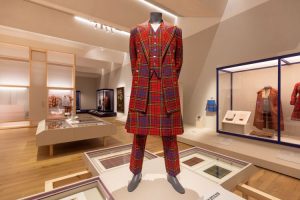 A red, blue and green tartan suit, comprising a lapelled jacket, waistcoat, kilt and trousers, stands in the centre. The background shows more tartan outfits and flat swatches shown in framed display cases.