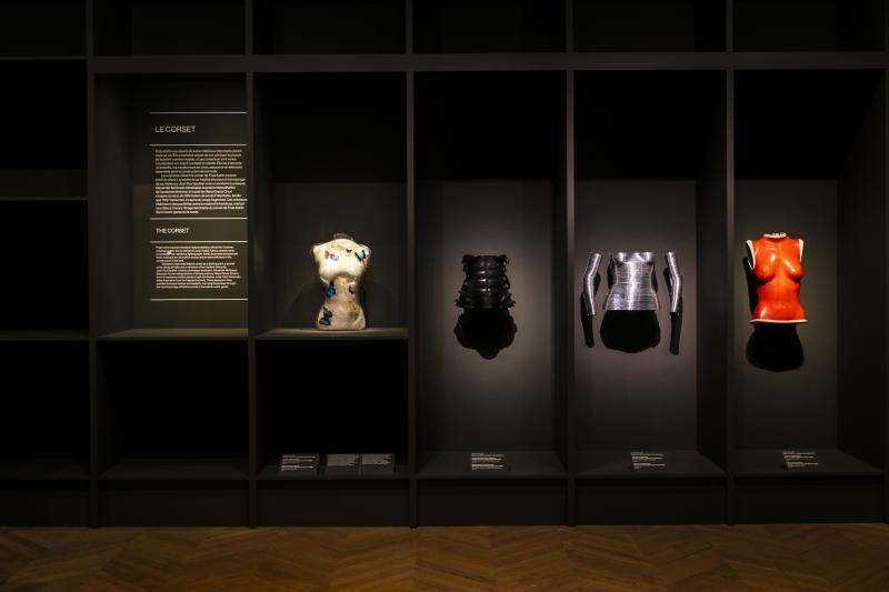 Four corsets softly lit displayed against a black wall. A panel of wall text is on the left side.