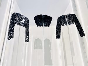 An exhibition view of three black lace bolero-style jackets are shown against a softly lit white backdrop.