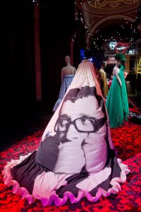 Exhibition view showing the back of a billowing ballgown with a monochrome printed photograph of Alber Elba looking away with his hands covering his mouth. More ballgowns are shown in the background along with fairy lights and neon hearts.