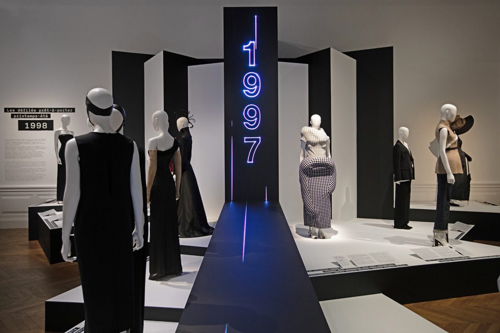 A number of white mannequins stand on monochrome plinths. In the centre is a black runway with '1997' in purple LED lights.