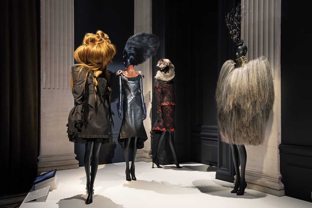 Four Alexander McQueen dresses are worn on black mannequins against a black and pillared background. Three mannequins have a feather or fur headpiece, with another wearing a voluminous ginger wig. The right-hand dress is a shaggy fur with stacked metal rings covering the neck. The second dress has a red tartan pattern covered with black lace. The third dress is black leather with an orange neckpiece with two bird heads resting over the shoulders. The fourth dress on the left is a puffed black minidress.