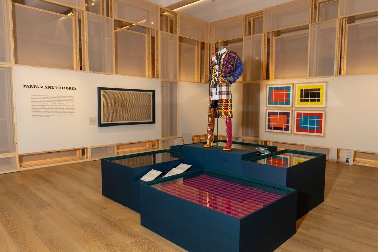 A multi-coloured tartan outfit and high heels is displayed atop a dark blue modular plinth. Text introduced by 'Tartan and the Grid' is shown on a wall alongside tartan swatches and patterns.