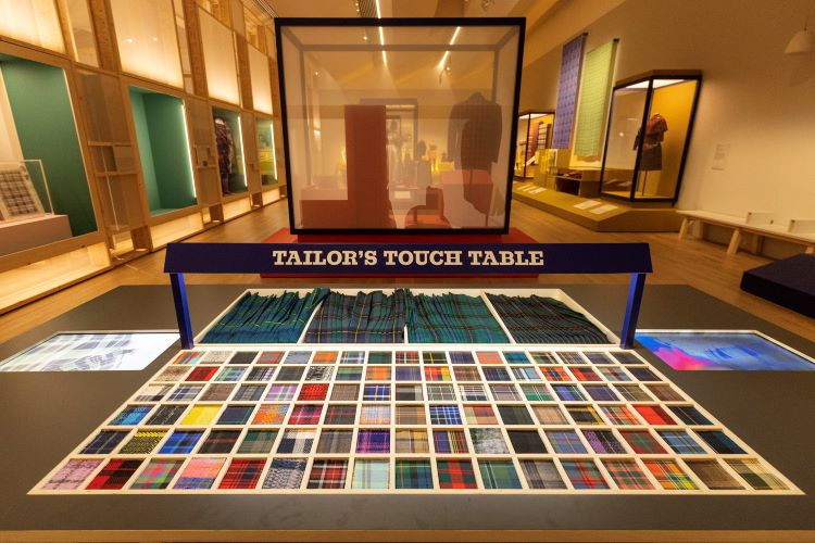 At the forefront shows 102 tartan swatches stationed in front of text saying 'Tailor's Touch Table'. Two digital screens are flat within the station. The background are tartan outfits displayed in modular frames.
