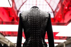 A close-up view of the back of a black tailored jacket, formed partly of crocodile skin,. In the background is the white lights and domed ceiling in red light.