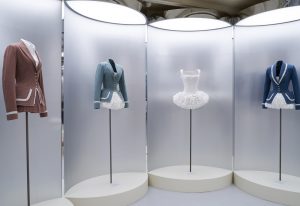 Four tailored outfits are displayed on black poles in front of translucent tubular backdrops. The far left is a brown riding-style jackets with white trim and a white shirt and brown pleated skirt. The other outfits include three white tutus; two of which are covered by blue riding-style jackets