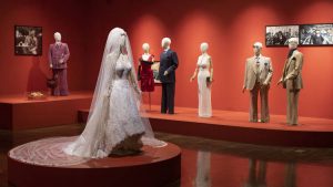 A veiled white wedding dress stands on a red circular plinth in the middle of the gallery. In the background are 1970s-styled suits and dresses, all worn on white mannequins, on plinths and in front of red walls.