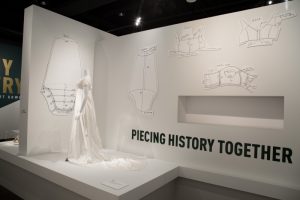 A white empress-style dress is worn on a white mannequin. Illustrated pattern cuttings are printed onto the wall behind the dress. The caption 'PIECING HISTORY TOGETHER' is printed near the bottom of the wall in large font.
