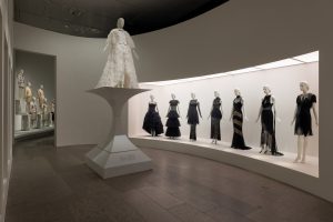 A mannequin wearing an ivory waistcoat, bow-tie and feathered coat stands atop a concave plinth. It overlooks a group of black dresses set inside an alcove on the right. In the background are more dresses staged on a pyramid formation.