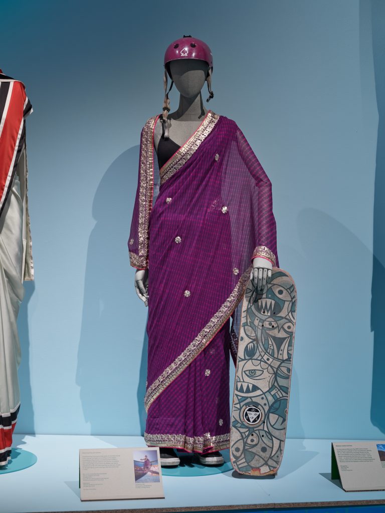 A purple chiffon sari has a silver border and worn on a mannequin. The mannequin holds a skateboard with cartoon faces and a purple helmet.