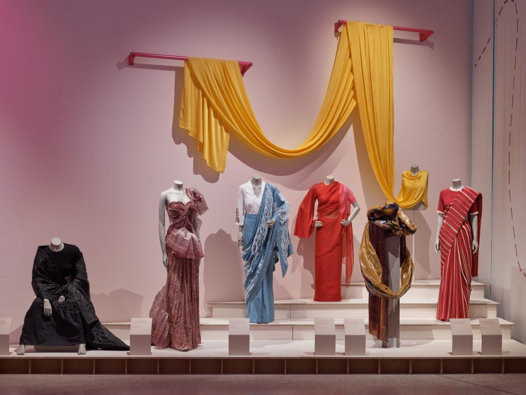 Seven saris are displayed on a white tiered plinths in a variety of poses. The sari at the back is made from an elongated piece of yellow fabric which is draped on two drawers above the display.