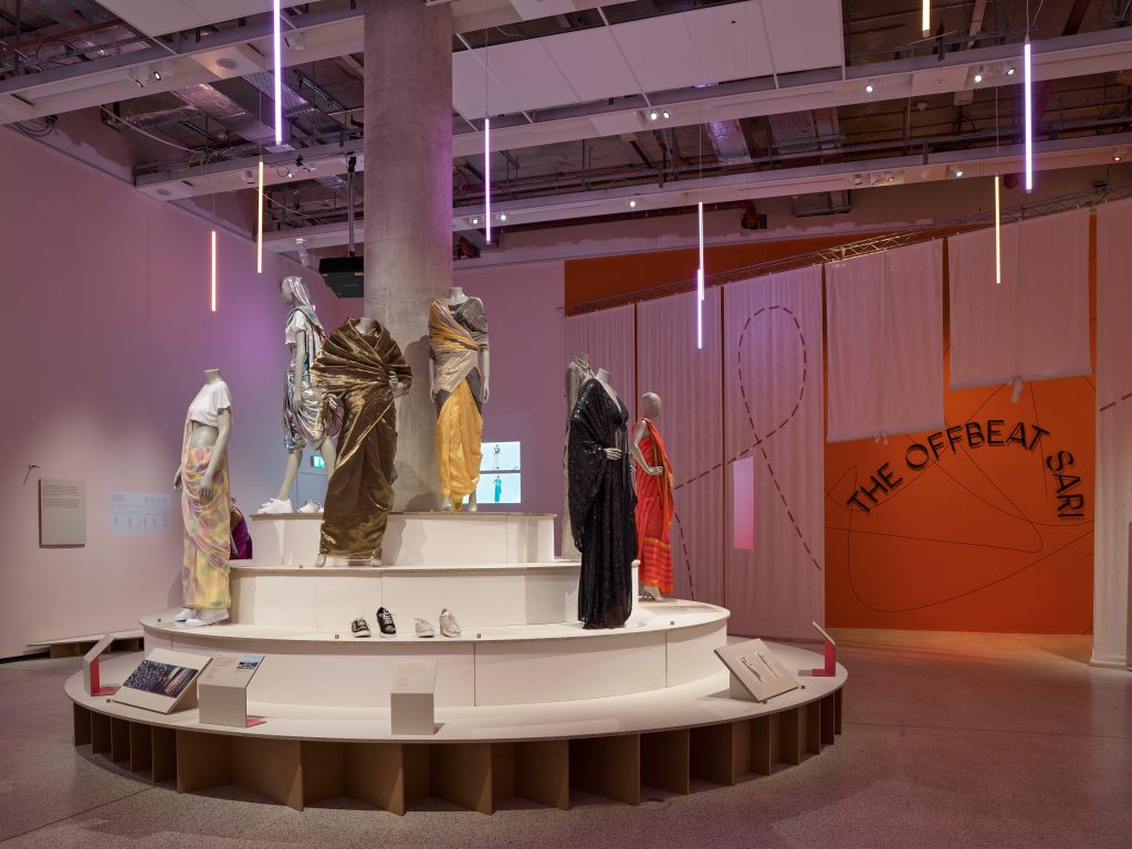 Six saris are displayed on mannequins, standing a tiered white circular plinth with a concrete spine. Vertical LED lights are hung from the ceiling overhead. 'THE OFFBEAT SARI' is printed on an orange wall in the background.