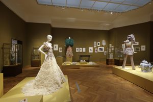 Gallery view showing three dressed mannequins on low yellow plinths, dissplayed alongside prints, vases, and display cases of objects.