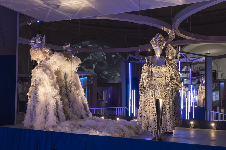 A pearl and jewel-encrusted silver gown, overcoat and papal mitre, worn by Rihanna originally, is displayed on a mannequin next to a silver and white feathered outfit, originally worn by Elton John.