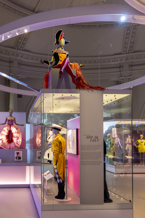 A mannequin wearing a blue and red, gold-edged Bicorn, Admiral coat and red buckled heels sits atop a display case. Beneath are more outfits including the back of a gold navy coat with sailor's hat and harlequin-patterned trousers, and Janelle Monae's Vulva trousers.