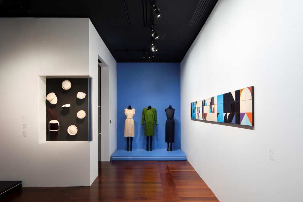 Gallery view showing white hats displayed against a black alcove set into a white wall. To its right are three dresses against a blue wall near an artwork of mosaic tiles.