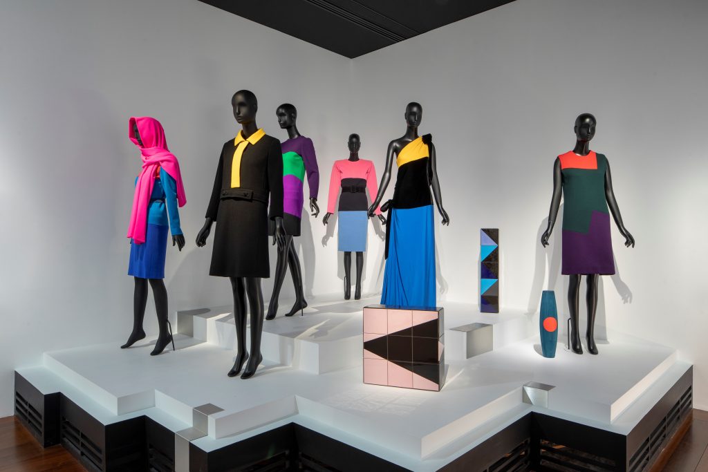 Six dresses in colour-blocked geometric patterns are worn on black mannequins alongside geometric artworks on a white plinth.