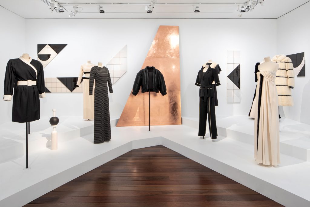 A range of monochrome outfits are displayed on tailor's dummies in front of monochrome mosaic artworks. In the centre of the background is a rose gold sloped structure.