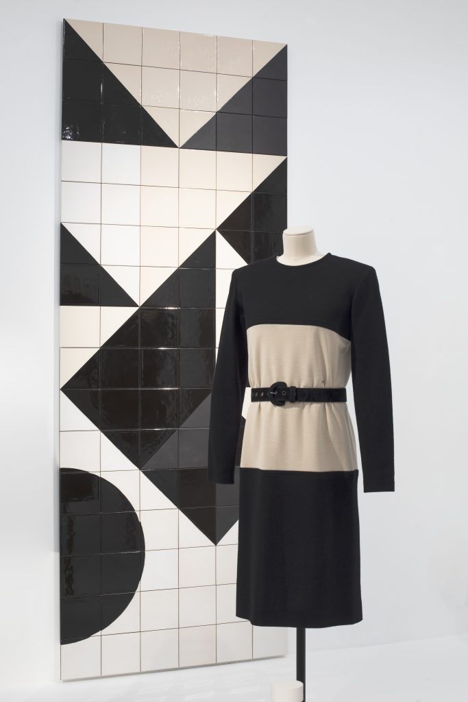 A monochrome long-sleeved dress is displayed on a tailor's dummy in front of a monochrome mosaic artwork.