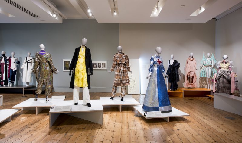 Gallery view of mannequins wearing a range of outfits on three sections. In the backleft are three mannequins; in the centre, four mannequins stand on individual white plinths; on the right, four mannequins stand on four brown wooden plinths.