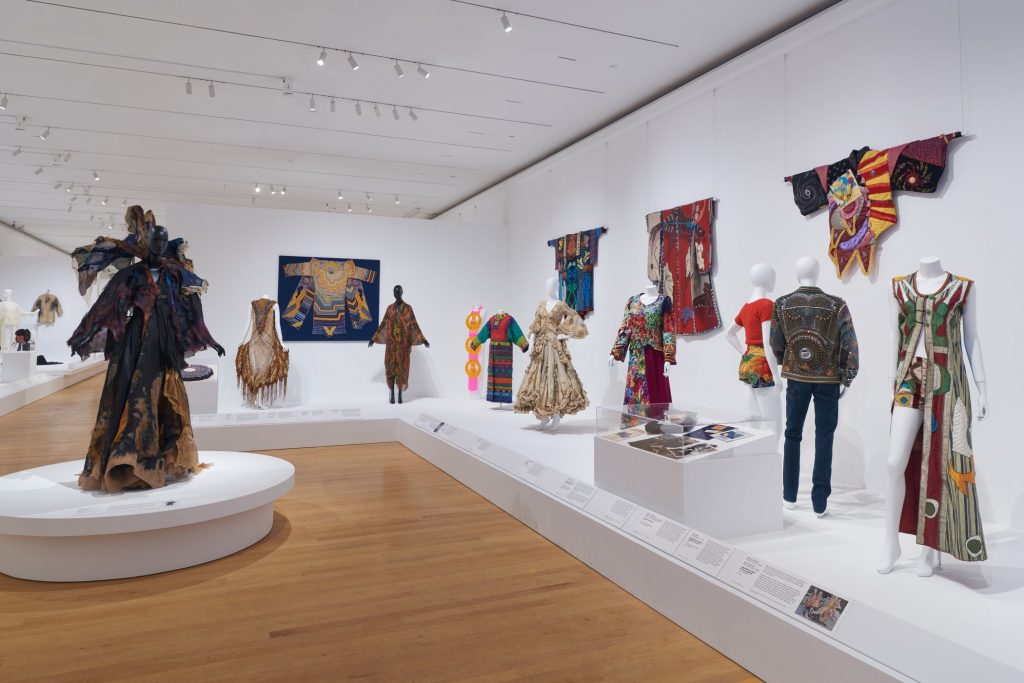 Gallery view showing clothes worn on mannequins and hung on walls. These are displayed on low-level white plinths with label strips and white walls.