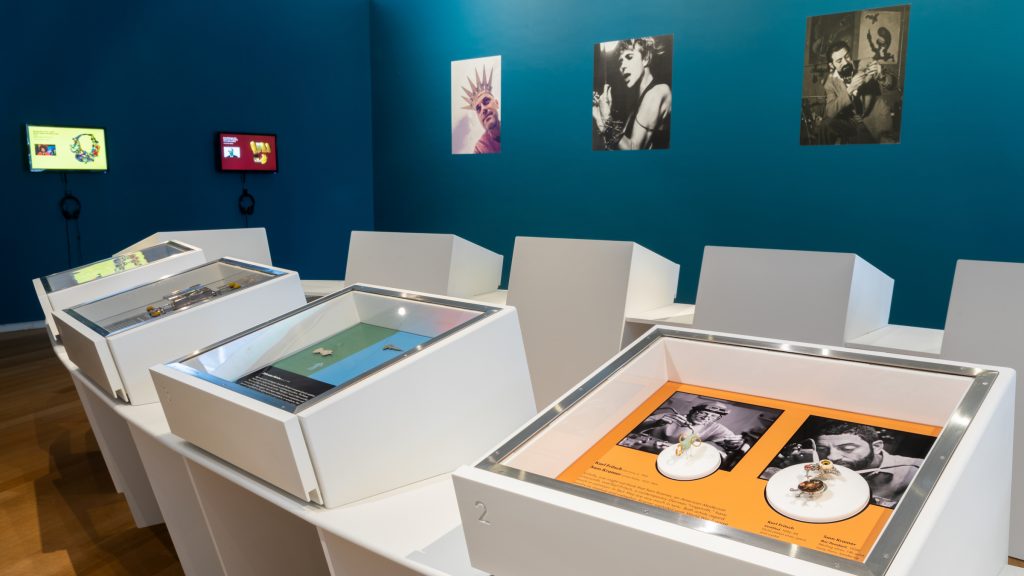View of the gallery. A long white plinth features nine shallow display cases, exhibiting pieces of jewellery. Images of men wearing jewellery are pasted onto a teal wall behind.