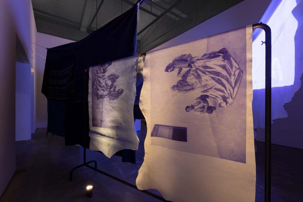 Textiles showing depictions of people holding cloth are draped on steel frames. In the background is a film projection.