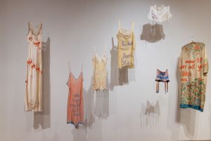 Six embroidered nightgowns and a pair of garters are hung near a white wall, creating shadows against the background.