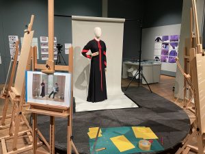 A mannequin wears a black gress with a red bow, collar and striped arms, standing in front of a screen as though on a photoshoot. It is surrounded by easels holding paintings and text.