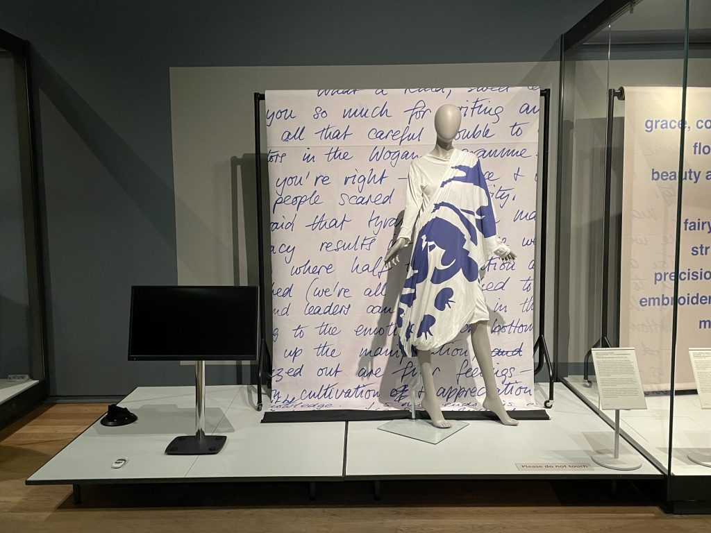 A mannequin wearing a white and blue graphic dress stands on display in front of a printed screen, hung by a rail. The print shows large hand-written text.