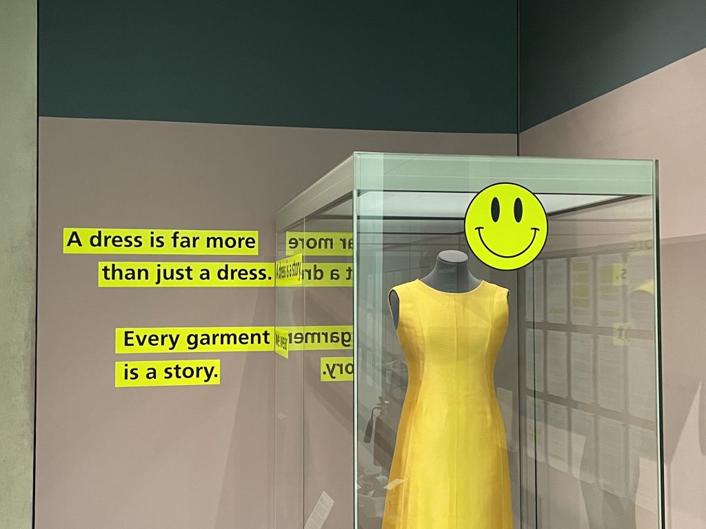 A golden-yellow sleeveless dress is displayed in a case, next to a smiley face design and black text on yellow. The text reads: 'A dress is far more than just a dress. A dress is a story. Every garment is a story.'