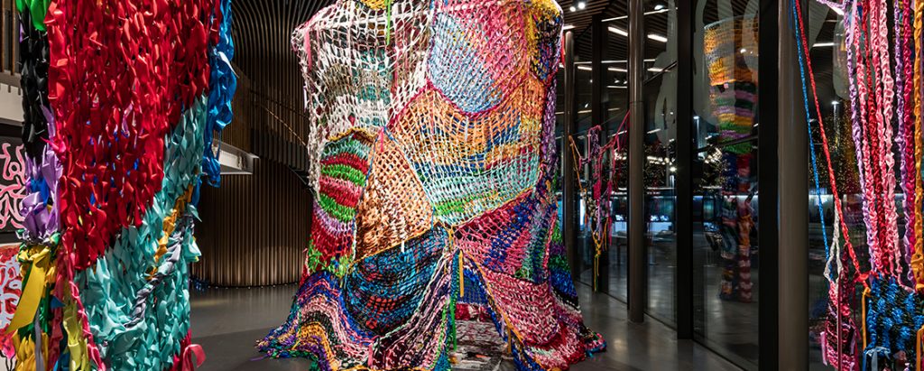 Gallery view showing a large, colourful knitted structure which visitors can sit inside, made of ribbons.