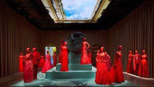 Gallery view showing a projection of a skylight. Underneath is a large, grey replica of a marble face. Surrounding it is a group of red Valentino dresses, worn on red mannequins.