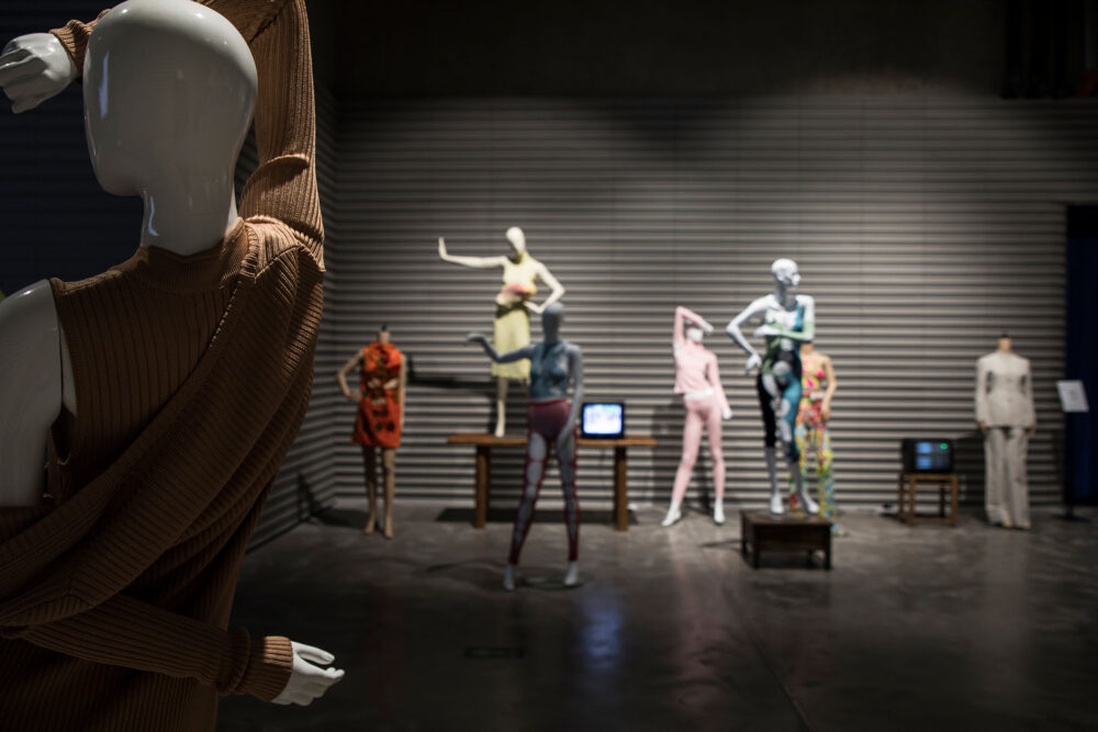 In the front left is a partial viewing of a mannequin wearing a jersey with its arm over its head. In the background are five other mannequins standing in different poses wearing brightly coloured clothes. A small TV screen sits in the middle.