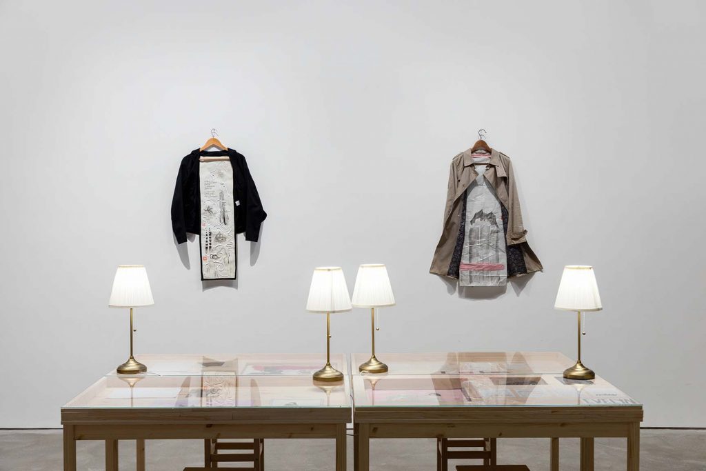 Two garments, a black hoody and beige trench coat, are hung on a white wall with wooden hangers and artwork. Four glass-topped tables featuring artwork and text are in front, with lamps on top.