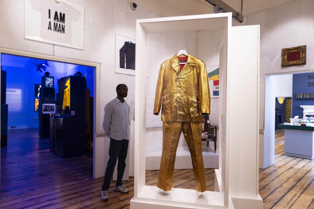 A gold metallic suic is hung in a white wooden display frame. In the background is a blue-lit gallery; above the doorframe is a T-shirt emblazoned with the phrase 'I AM A MAN'.