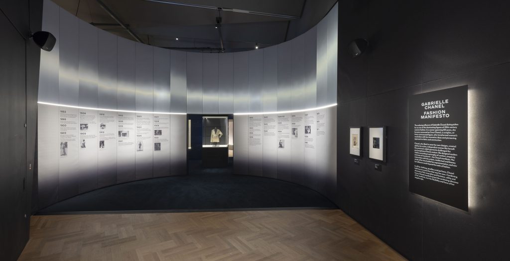Gallery view showing a timeline on a curved grey wall. In the centre is a cream top in a display case. On the right-hand side are two photographs and a large label entitled 'Gabrielle Chanel: Fashion Manifesto',